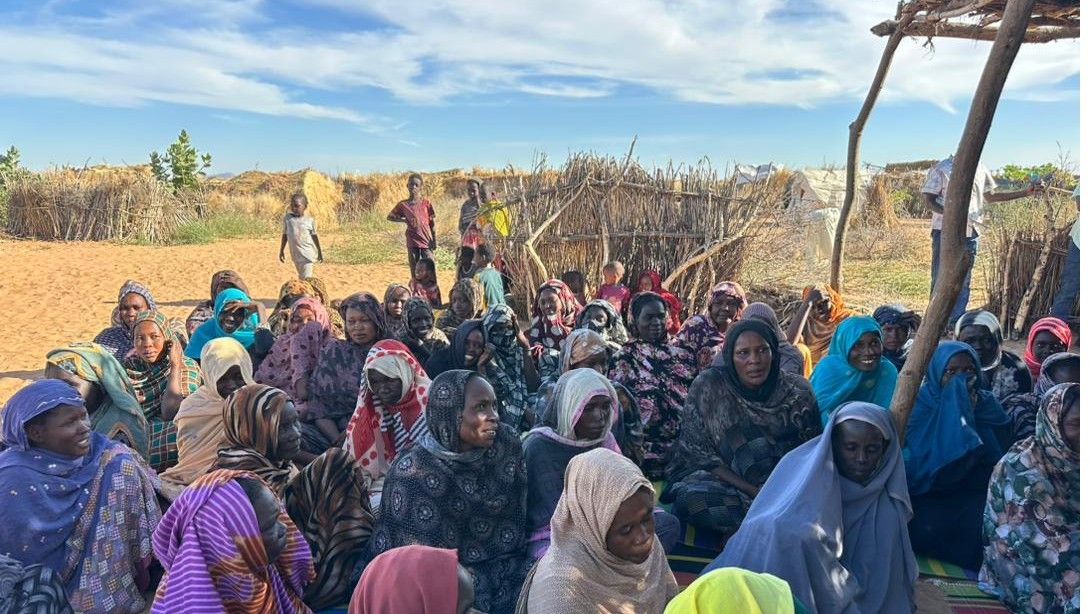 Women seated together on woven mats under an open sky at a camp gathering
