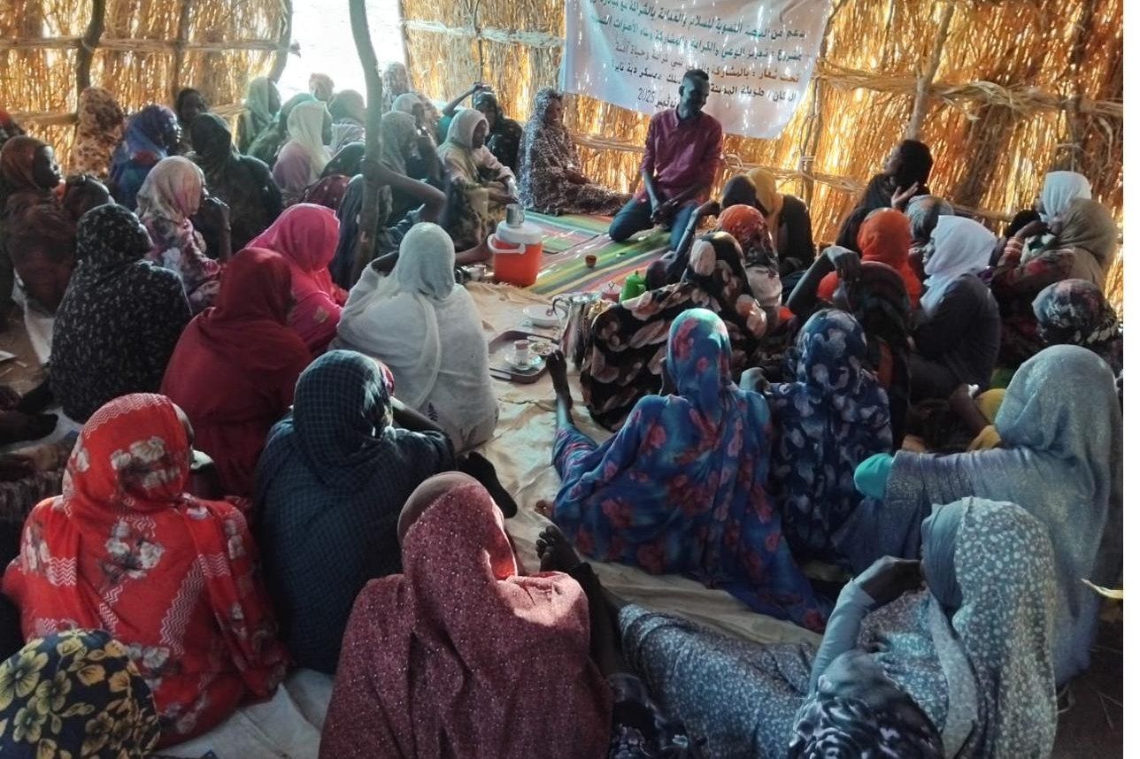 A large gathering of women seated on mats beneath a straw shelter