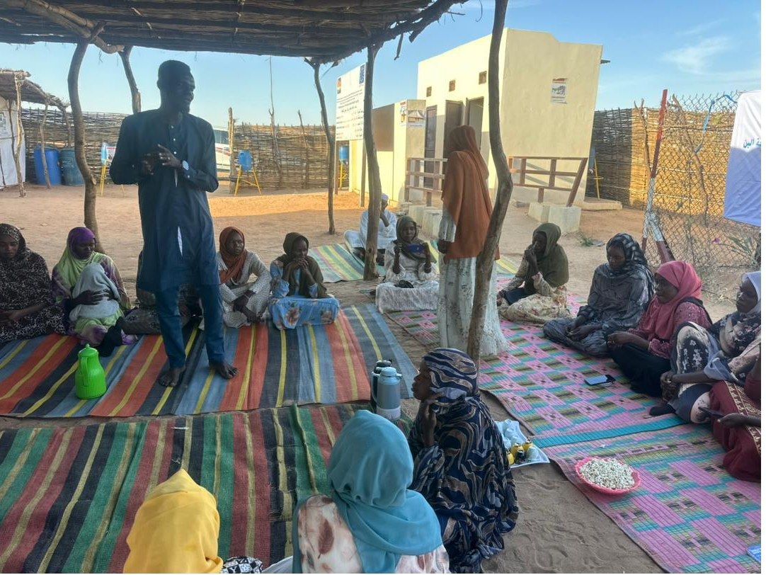 A facilitator standing to address women seated on mats under a shaded shelter